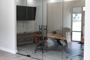 Glass-walled conference room with a long wooden table and brown rolling chairs; striped wall panels and a mounted screen reflect in the glass.