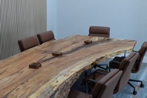 Large rustic wooden conference table with a natural edge, surrounded by brown cushioned chairs in a modern meeting room; small wooden boxes sit on the table.