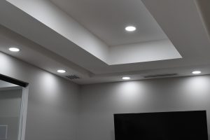 Modern interior ceiling with layered rectangular beams and circular recessed lights; a mounted flat-screen TV is visible on the right.