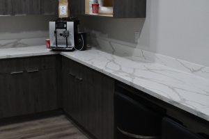 Coffee station on a marble countertop with an espresso machine, cups, and a brown bag nearby in a dark-kitchen setting.
