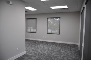 Empty office room with gray walls, two windows with blinds, and a patterned gray carpet.