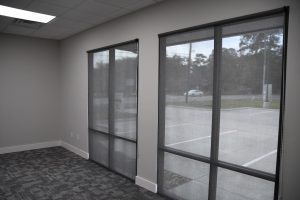 Office interior with tall glass doors and dark roller blinds; outside parking lot and trees visible.