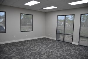 Empty modern office interior with large windows on two walls, gray walls, and a patterned gray carpet; ceiling fluorescent lights illuminate the space.