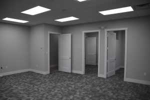 Office interior hallway with multiple white paneled doors along a gray wall and patterned carpet, bright fluorescent ceiling lights above