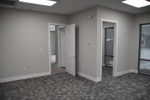 Empty office space with open white doors, gray walls, and carpeted floor; large windows on the right.