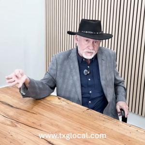 Man in a gray blazer and black hat sits at a wooden table during a video call or recording, with a striped wall behind and a watermark at the bottom edge.