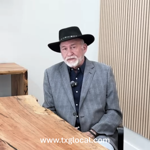 Older man in a gray blazer and black studded hat seated at a wooden table in a room with light walls and wood paneling, watermark visible at bottom.